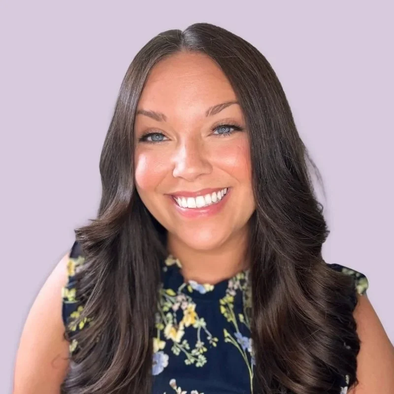 A woman with long, dark brown hair wearing a sleeveless navy blue dress with yellow and white floral patterns, smiling against a light purple background.