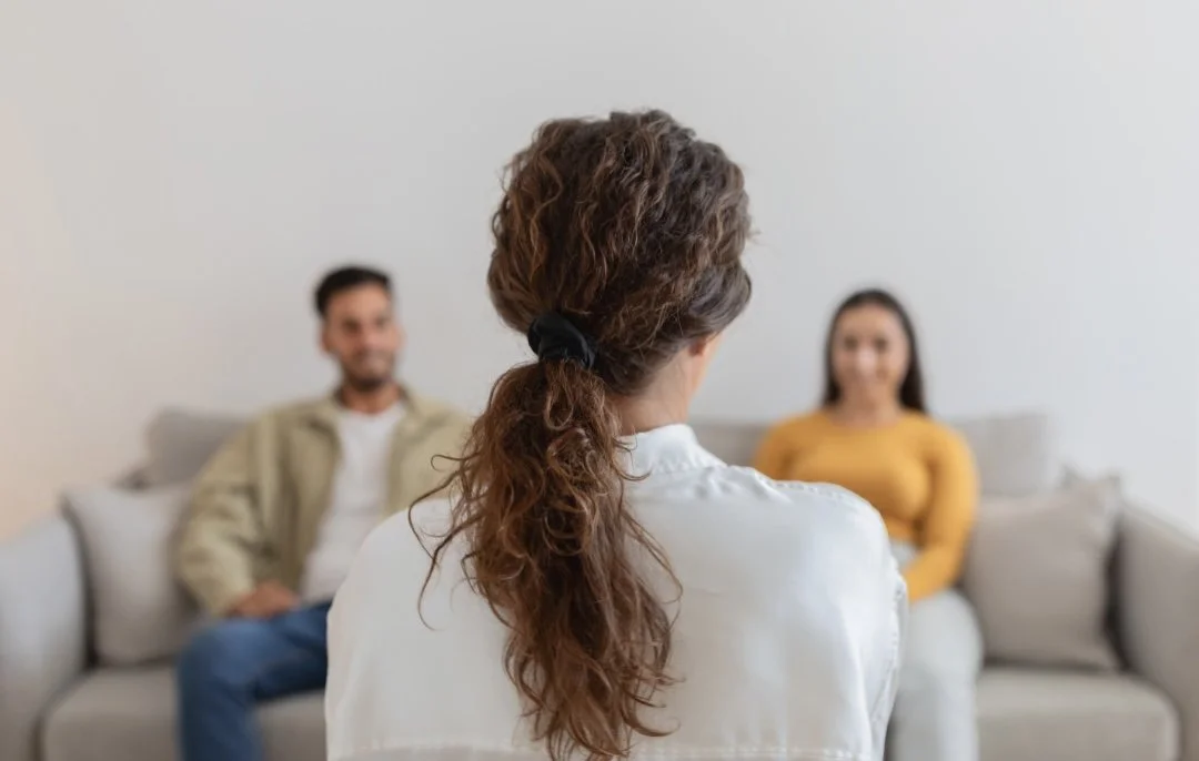 A woman with curly hair tied with a black scrunchie facing away, speaking to a couple sitting on a sofa during a therapy session.