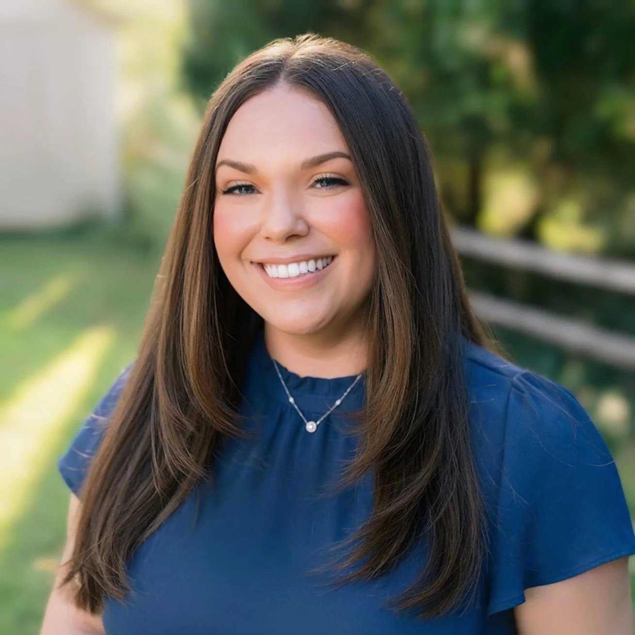 A woman with long brown hair, smiling, wearing a blue top and a silver necklace, standing outdoors with greenery and a wooden fence in the background.