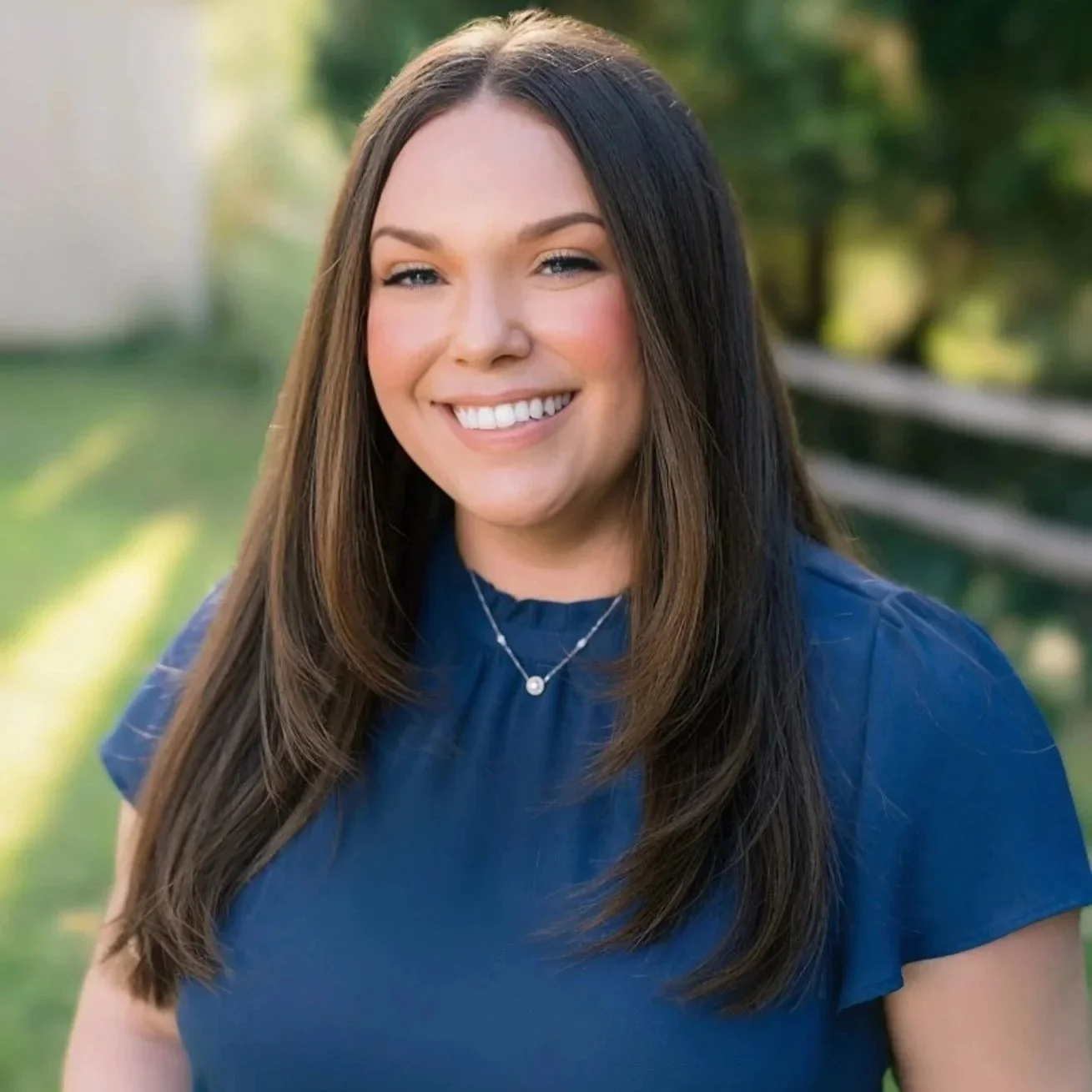A woman with long brown hair smiling outdoors, wearing a navy blue shirt and a delicate necklace, with a background of greenery and a wooden fence.