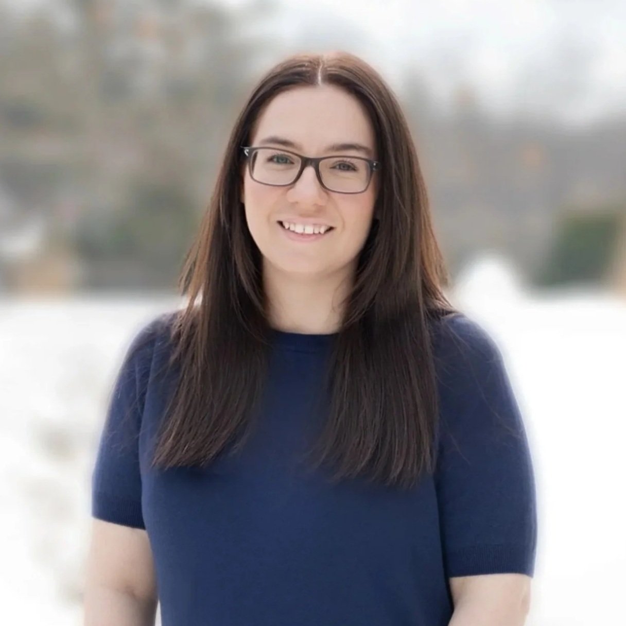 Headshot of a smiling woman with shoulder-length blonde hair, wearing hoop earrings and a white blouse with ruffles on the front, against a blurred beige background.