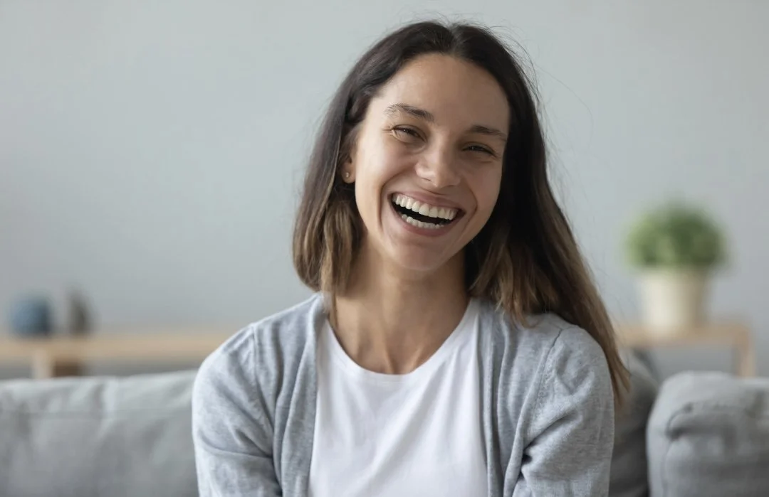A woman with shoulder-length brown hair smiling and laughing while sitting on a couch in a bright living room.