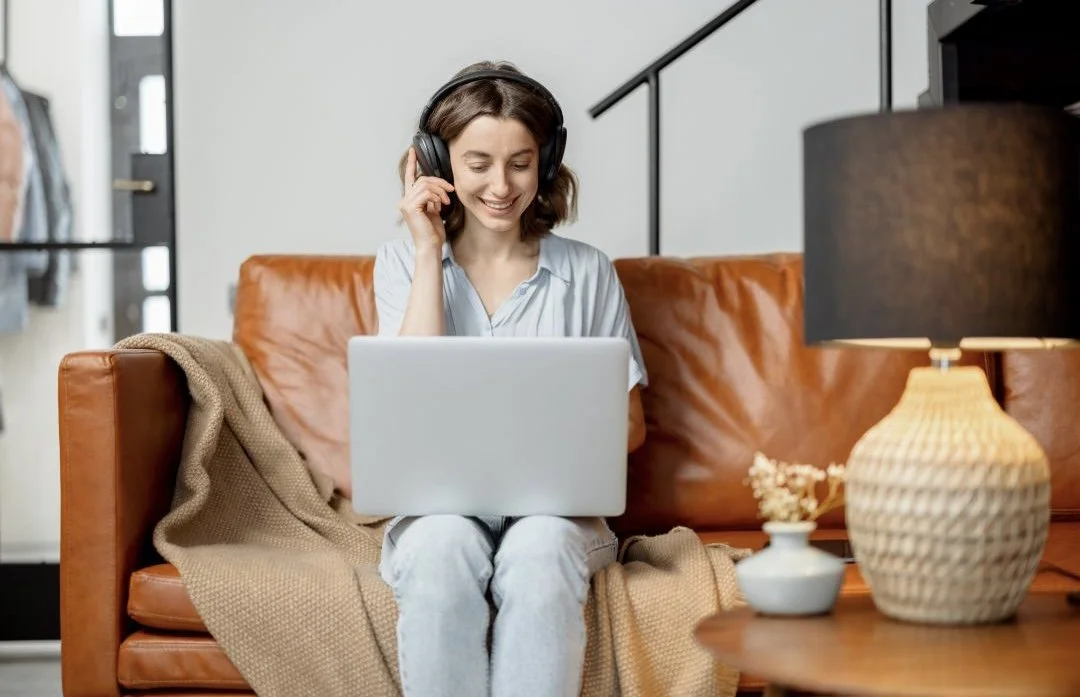 A woman sitting on a brown leather sofa, wearing headphones, looking at a laptop and smiling, with a beige blanket on her lap, in a cozy living room.