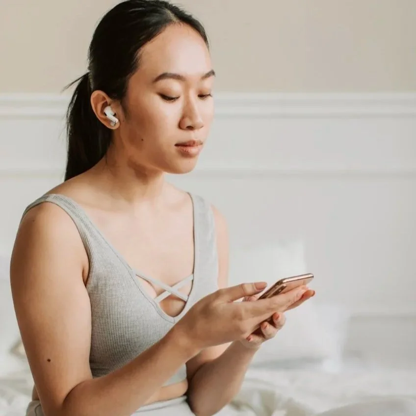 Young woman with dark hair in a ponytail, wearing a gray tank top and wireless earbuds, looking at her phone.