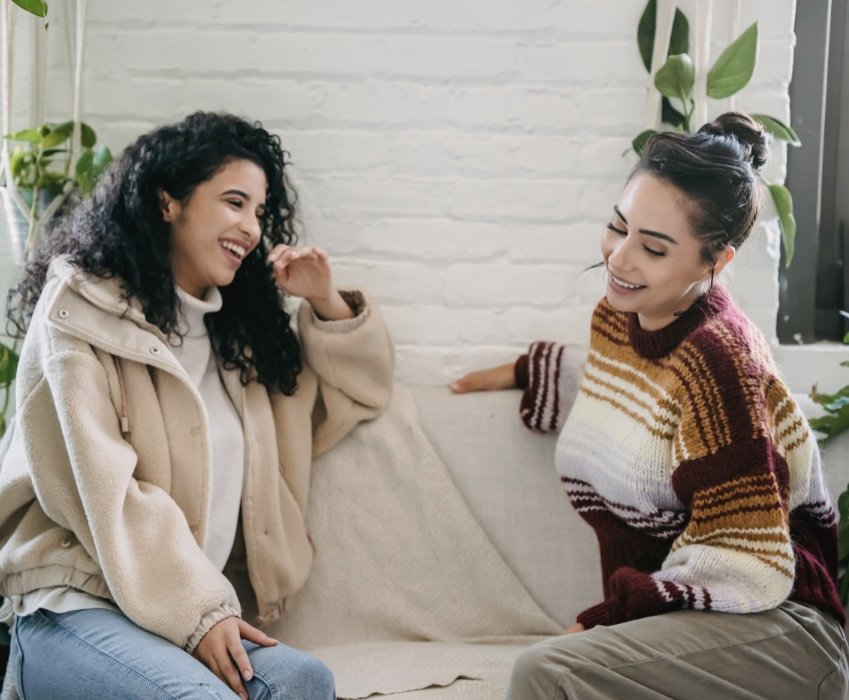 Two women smiling and laughing while sitting on a beige sofa in a bright room with white brick wall and green plants.