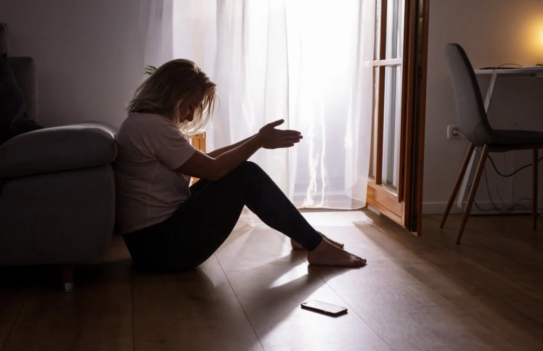A woman sitting on the floor next to a couch with her head bowed and hands raised, in a room with sunlight coming through a sheer curtain, and a smartphone lying on the wooden floor nearby.