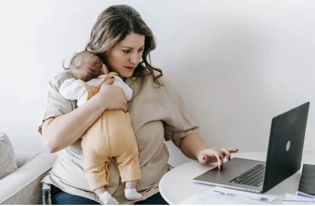 A woman sitting at a table working on a laptop while holding a baby on her shoulder, with the baby’s legs resting on her lap.