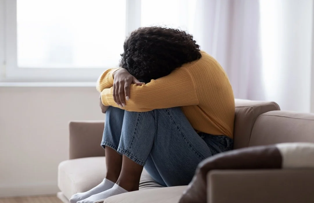 Woman sitting on a sofa, hugging her knees with her head down, appearing upset or distressed in a bright room with a window.