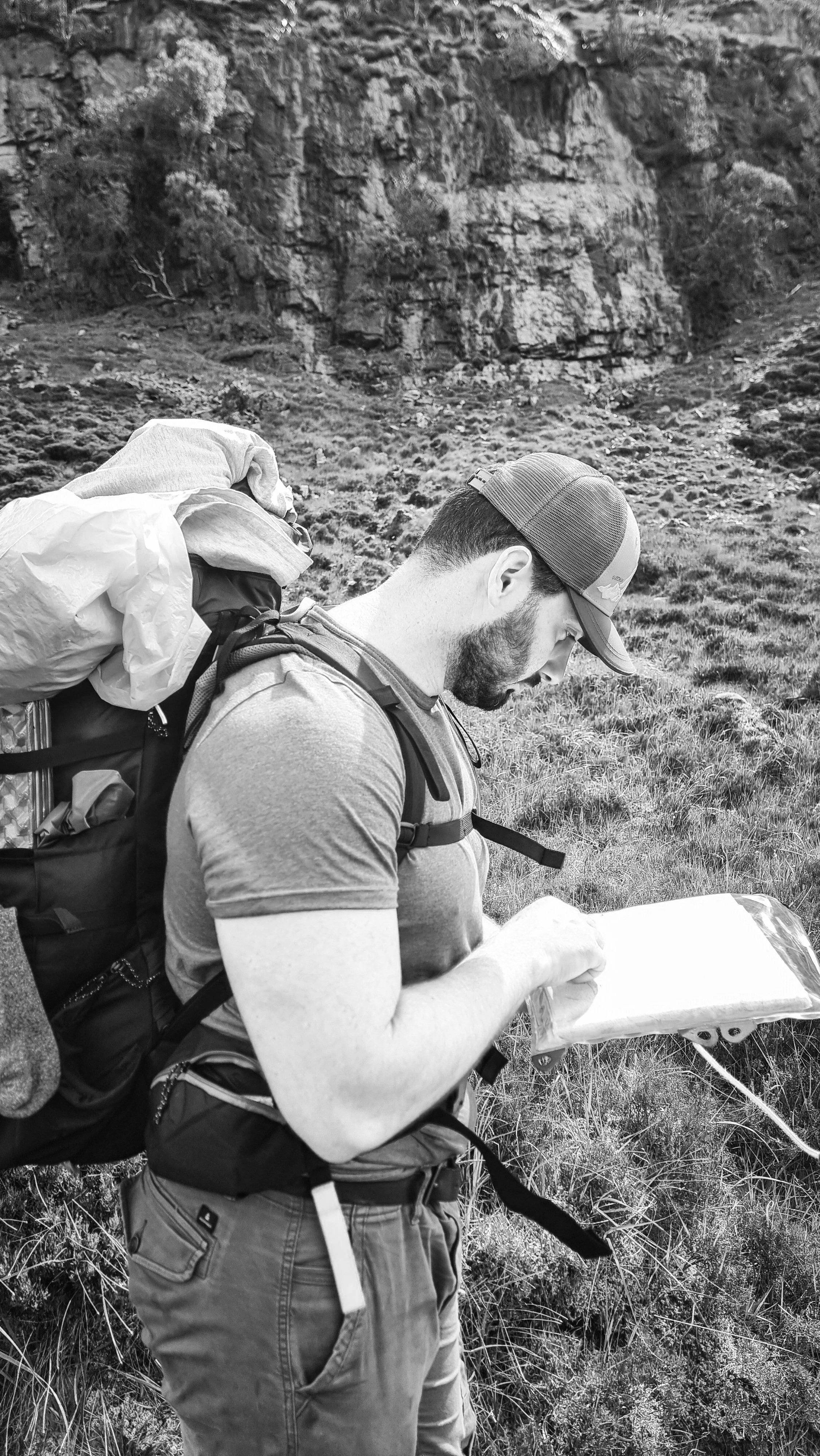 A man with a beard wearing a baseball cap, a t-shirt, and a backpack is standing outdoors in a grassy area, reading a map.