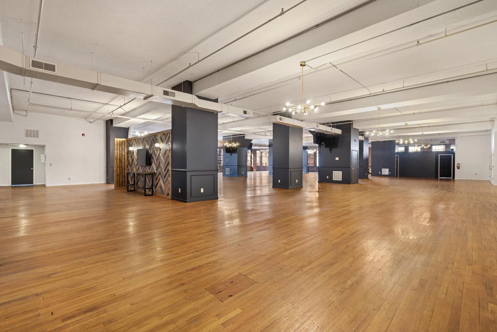 Monarch on Main’s expansive event hall with hardwood floors, white ceilings and exposed pipes, dark navy support columns, and bright natural light streaming through large brick-framed windows.