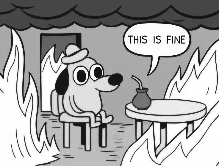 A cartoon dog sitting calmly in a room on fire, with flames and smoke around, stating 'This is fine' from a conversation bubble, with a table and a burning coffee pot nearby.