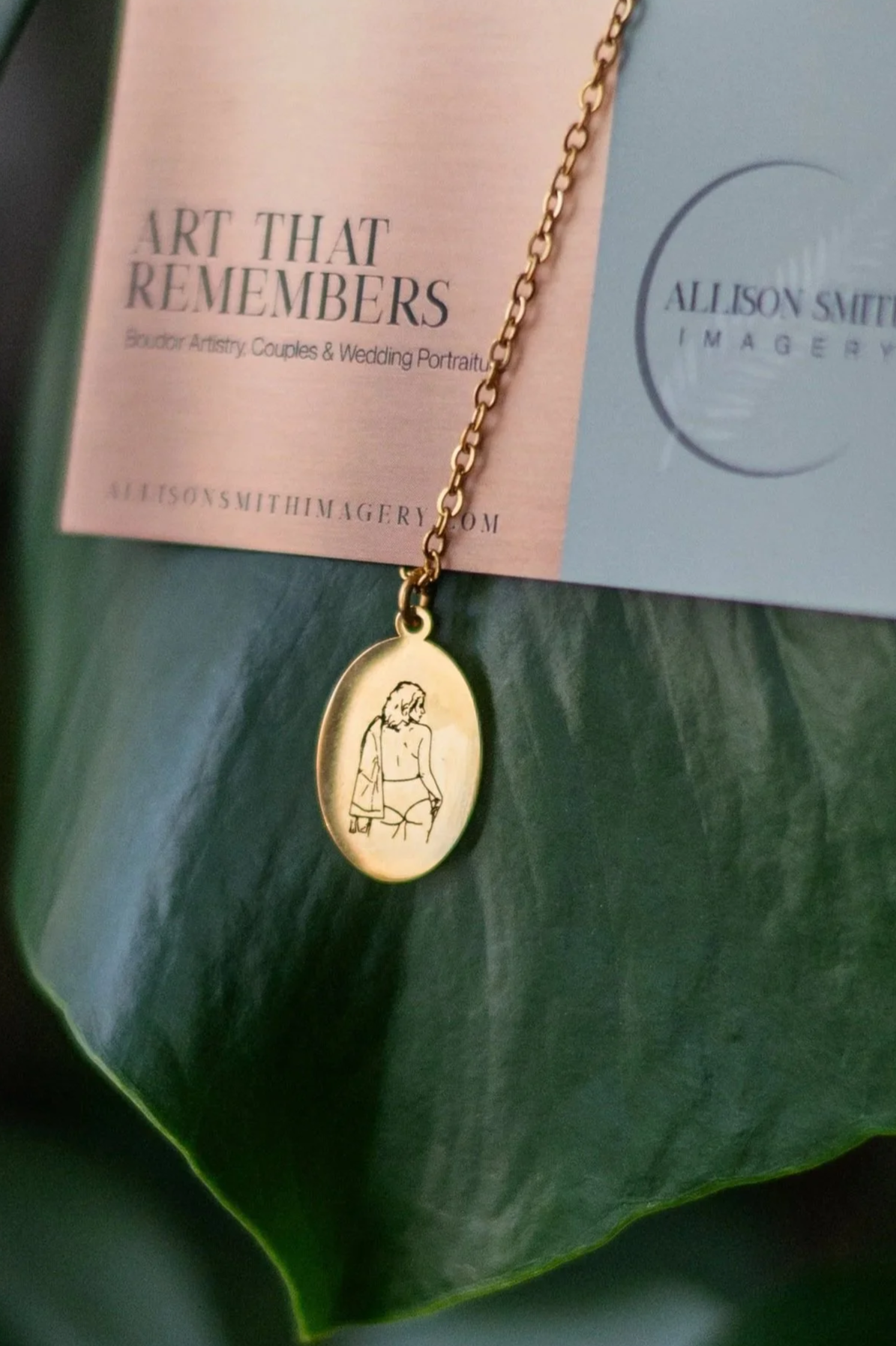 Gold necklace with an oval pendant featuring a minimalist line drawing of a woman in lingerie, resting on a green leaf with promotional cards in the background.