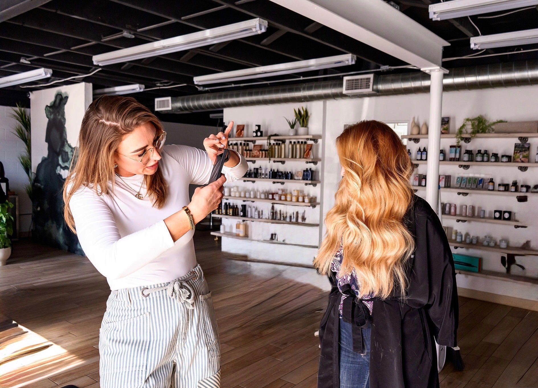 Two women in a store, one taking a photo of the other with a smartphone. Shelves with bottles and jars on the background.