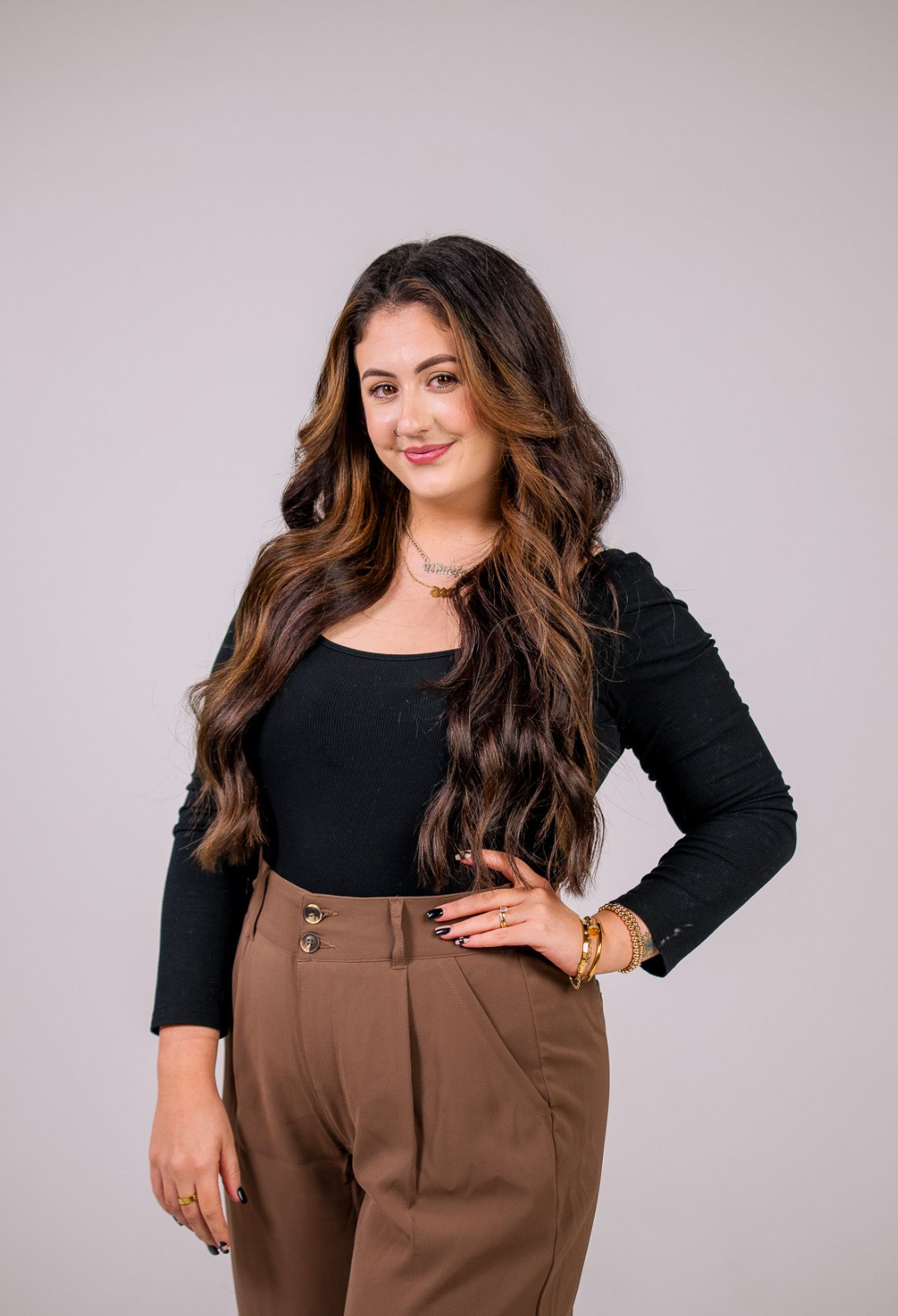 A young woman with long wavy brown hair, wearing a black long-sleeve top and tan high-waist pants, poses with her hand on her hip against a plain white background.