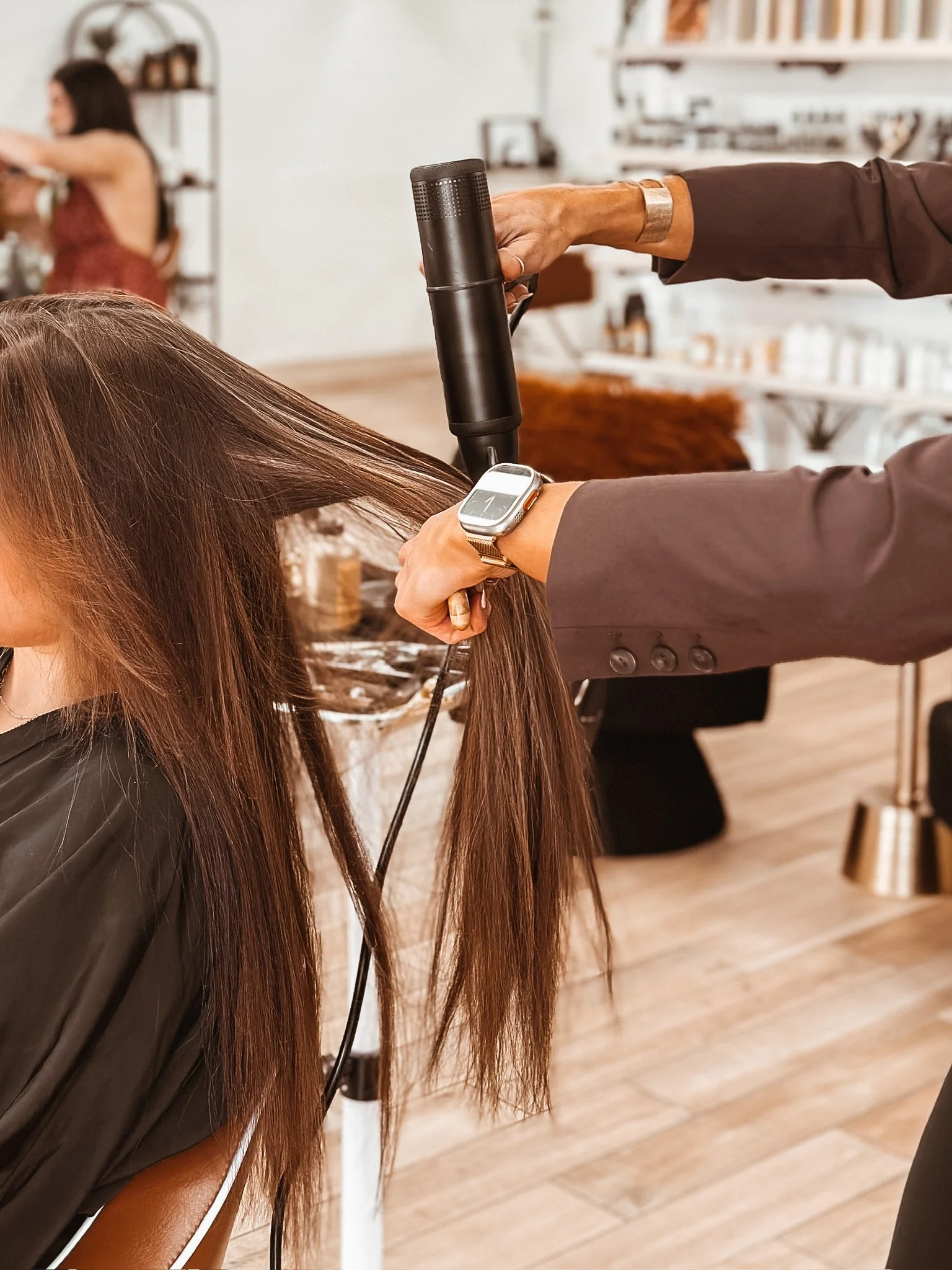 A person is drying a woman's long brown hair with a handheld blow dryer in a salon. The woman is seated, wearing a black cape, and the stylist is wearing a watch and a brown blazer.