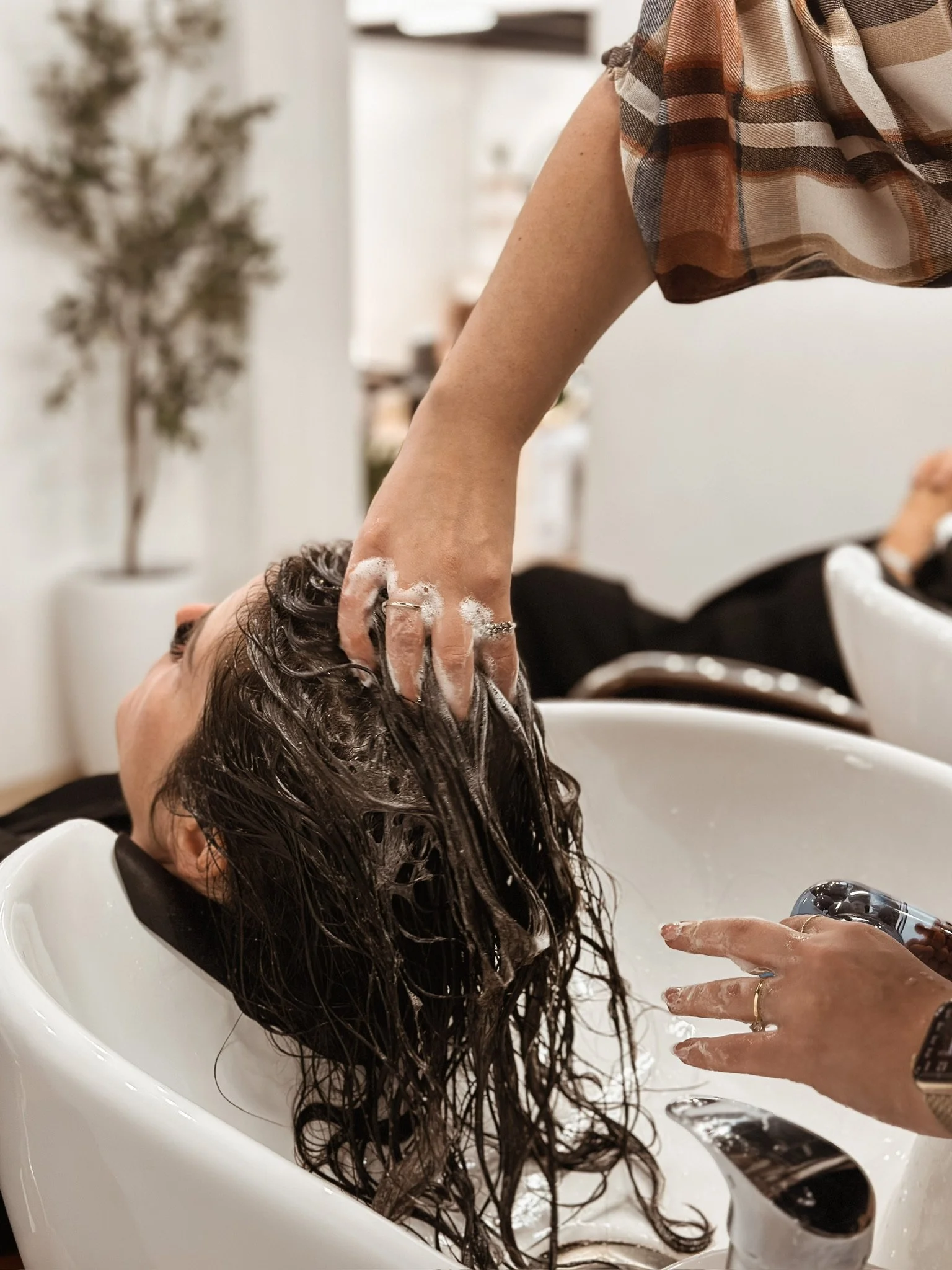 A woman is lying back in a salon sink with her head tilted backwards while a stylist washes her long, curly hair. The stylist has soap suds on their hand and is rinsing the woman's hair under a faucet.