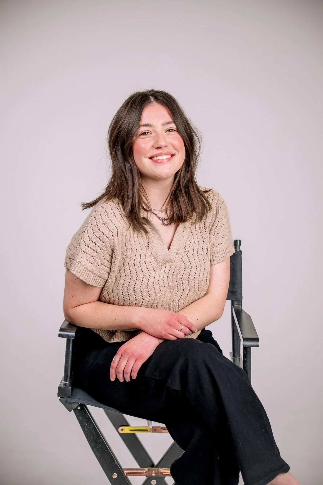 A young woman with shoulder-length brown hair, wearing a beige knit top and black pants, sitting on a director's chair and smiling at the camera against a plain light-colored background.