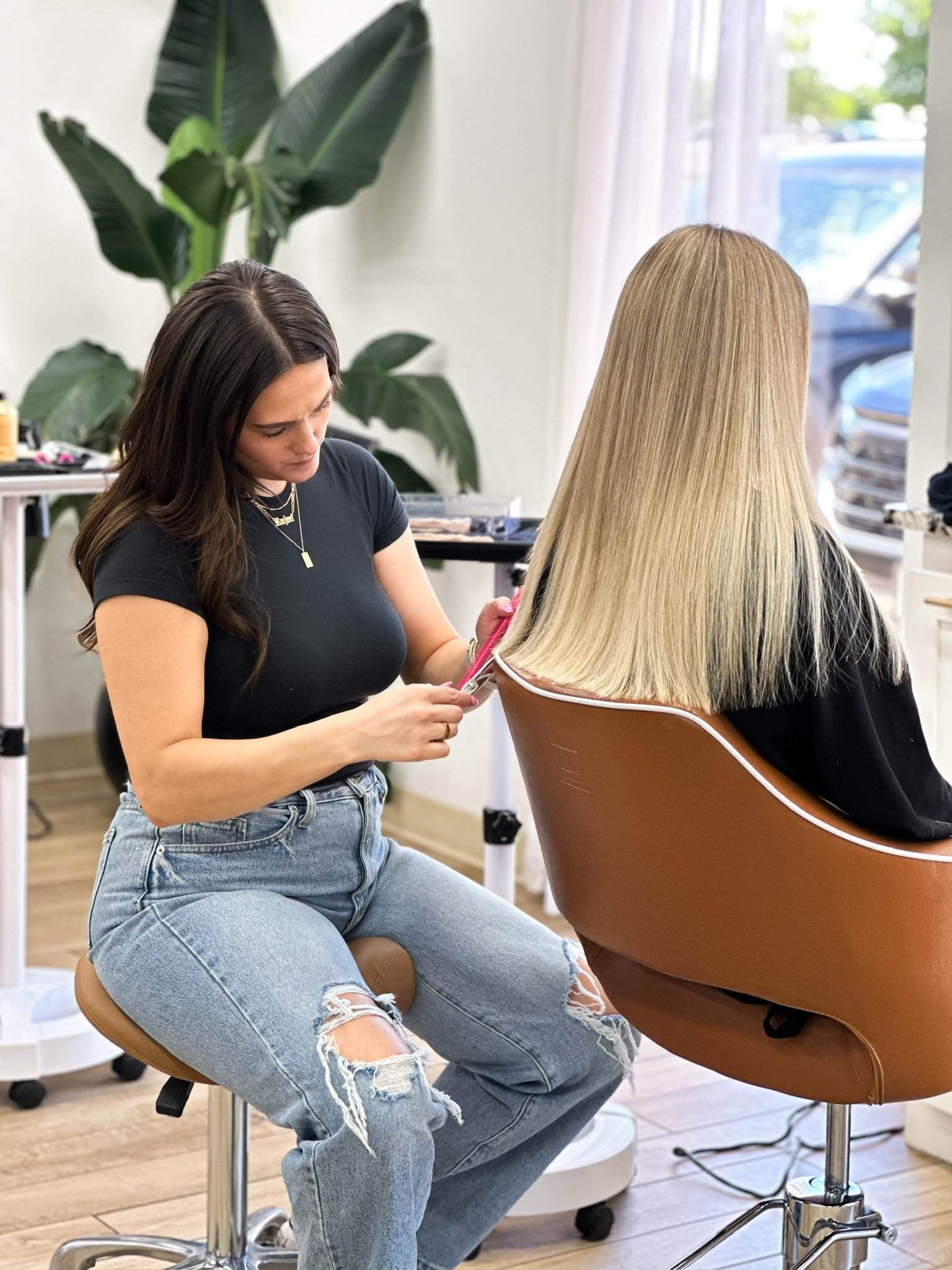 A hairstylist is cutting a woman's long blonde hair in a salon, seated on a brown chair with a large green plant and a window in the background.