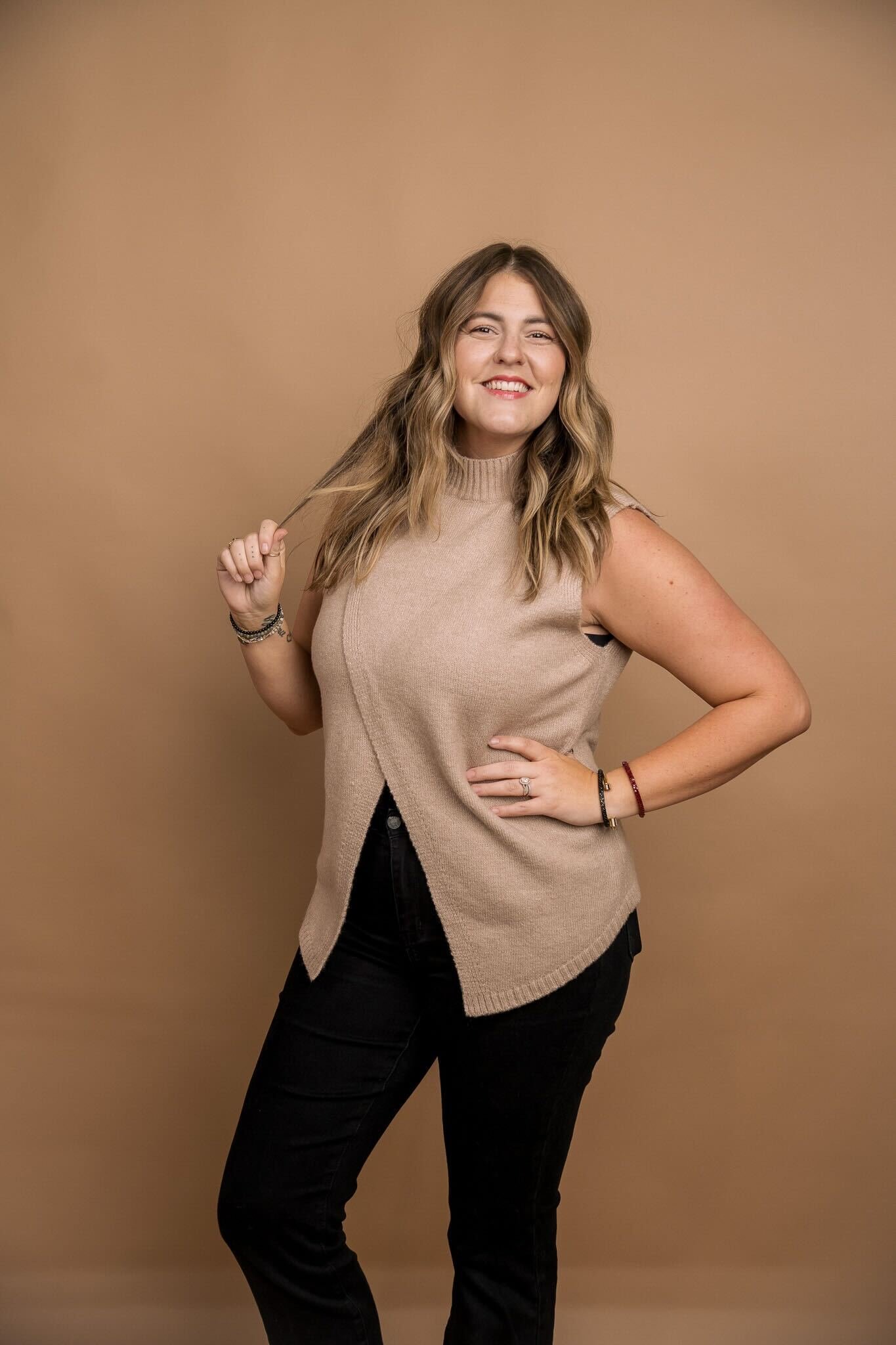 A woman with wavy, shoulder-length hair, wearing a sleeveless beige sweater and black pants, smiling and posing against a plain beige background.