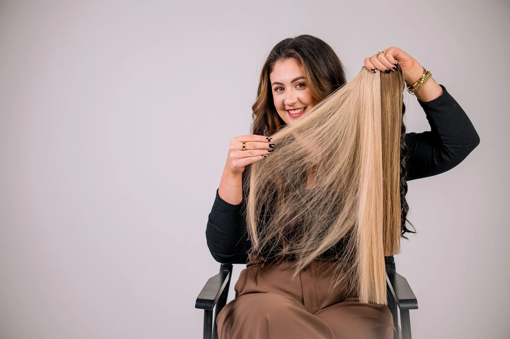 A woman with brown hair and a black long-sleeve shirt sitting on a chair, holding a large lock of blonde hair, smiling at the camera.