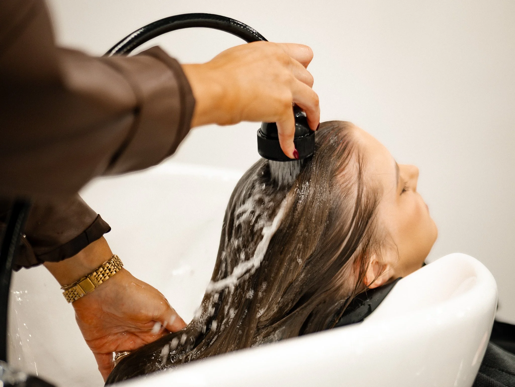 Person washing a woman's hair at a salon, with water running from a handheld showerhead as the woman reclines with eyes closed.