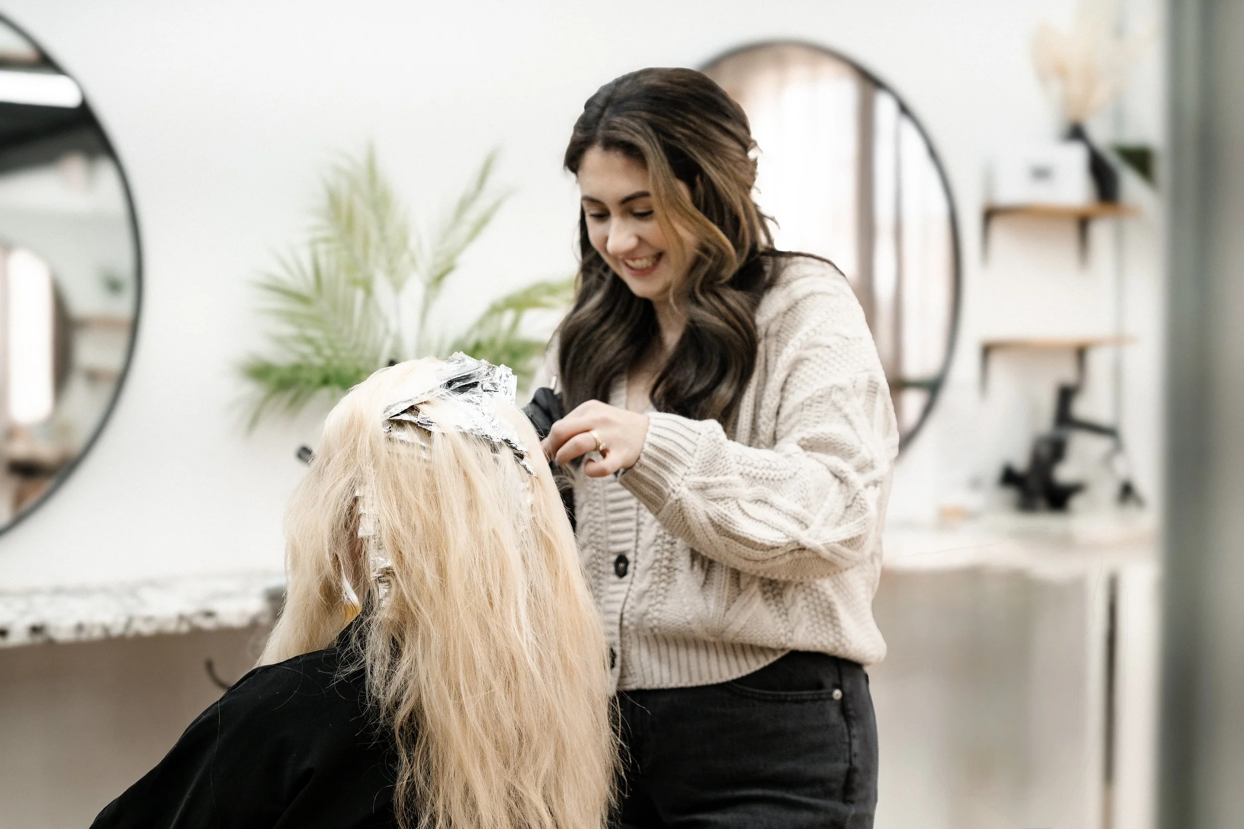 A hair stylist applying hair dye or toner to a client's blonde hair in a salon with mirrors and plants.