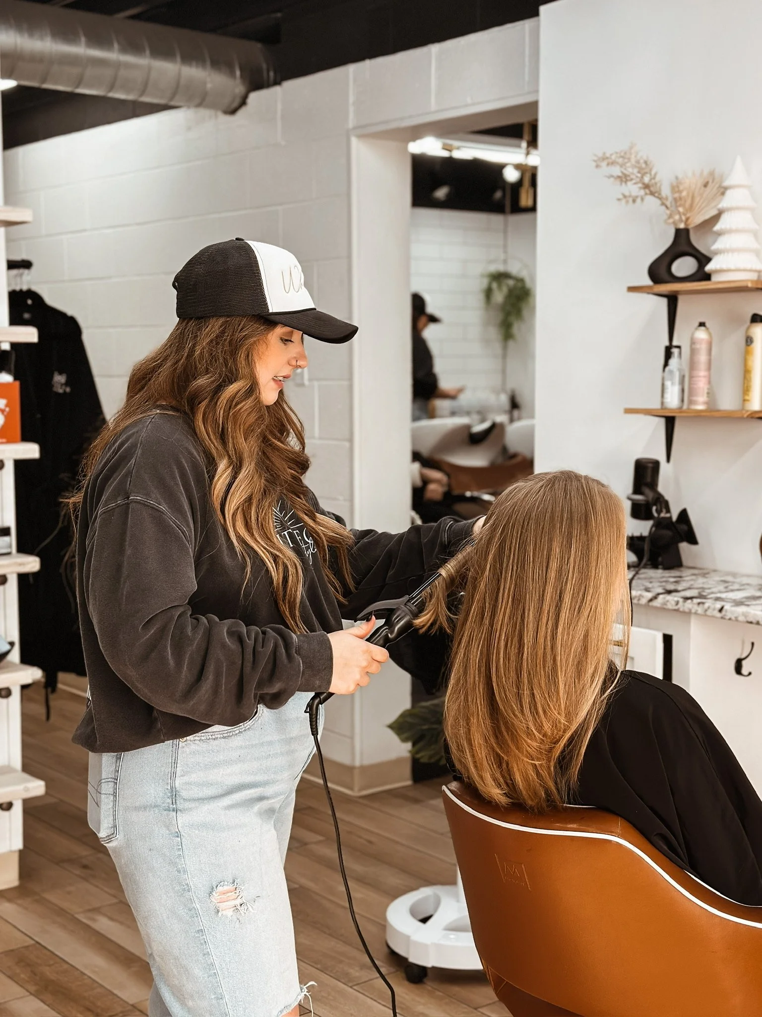 A woman with long wavy hair and casual attire styles a customer's hair in a salon, using a curling iron. The salon has a modern decor with wooden accents and decorative vases on shelves.
