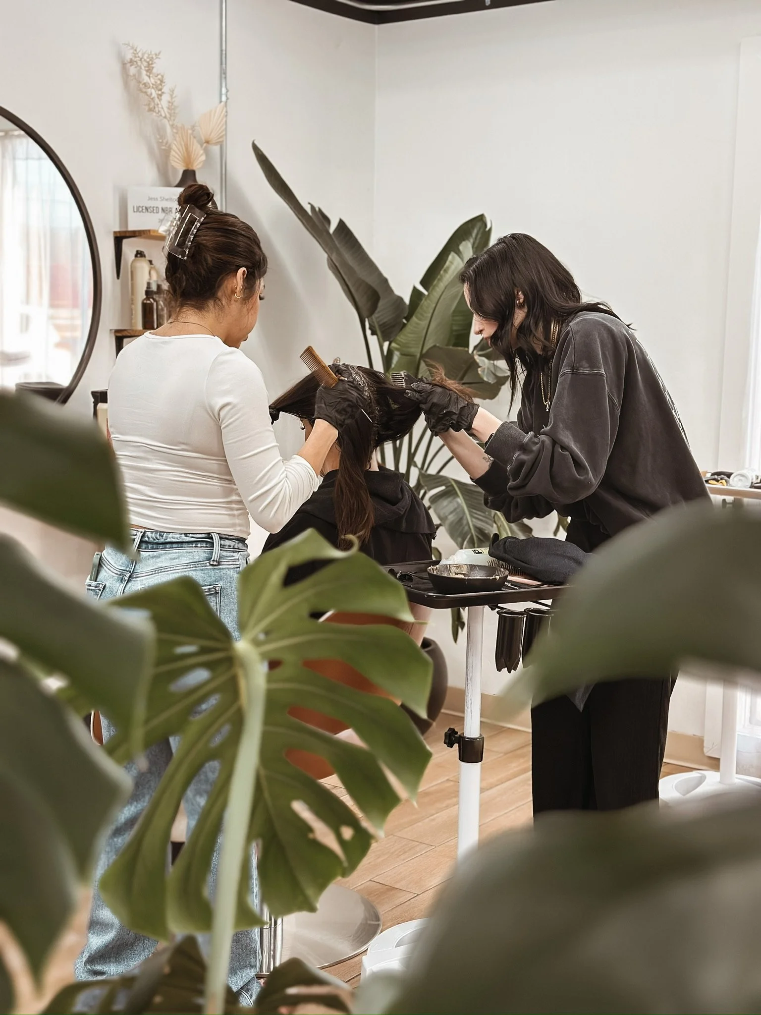 Two hairdressers working on a client's hair in a salon, with large green plants and wooden flooring visible.