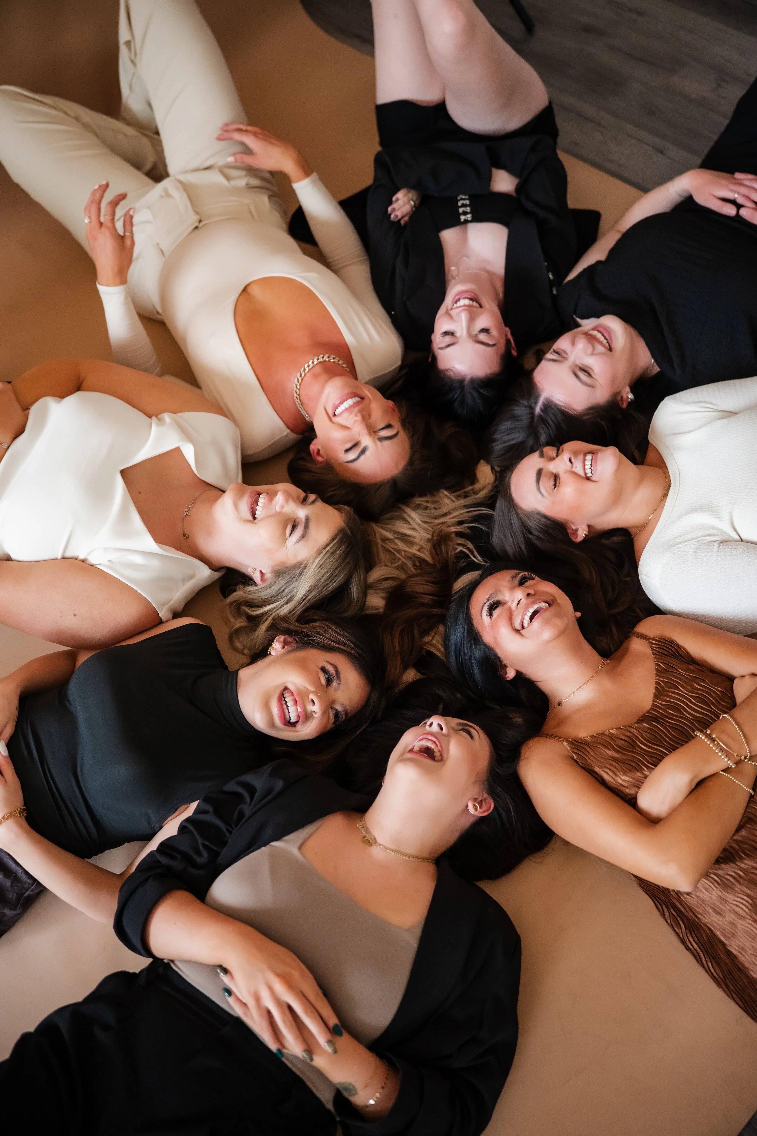 A group of nine women lying on the floor in a circle, smiling and laughing, dressed in various stylish outfits, with their heads close together.