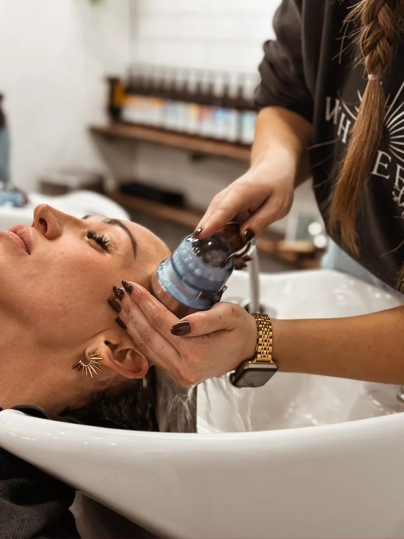 A woman getting her hair washed at a salon or spa, lying back with her eyes closed while a stylist or esthetician applies a treatment to her hair.