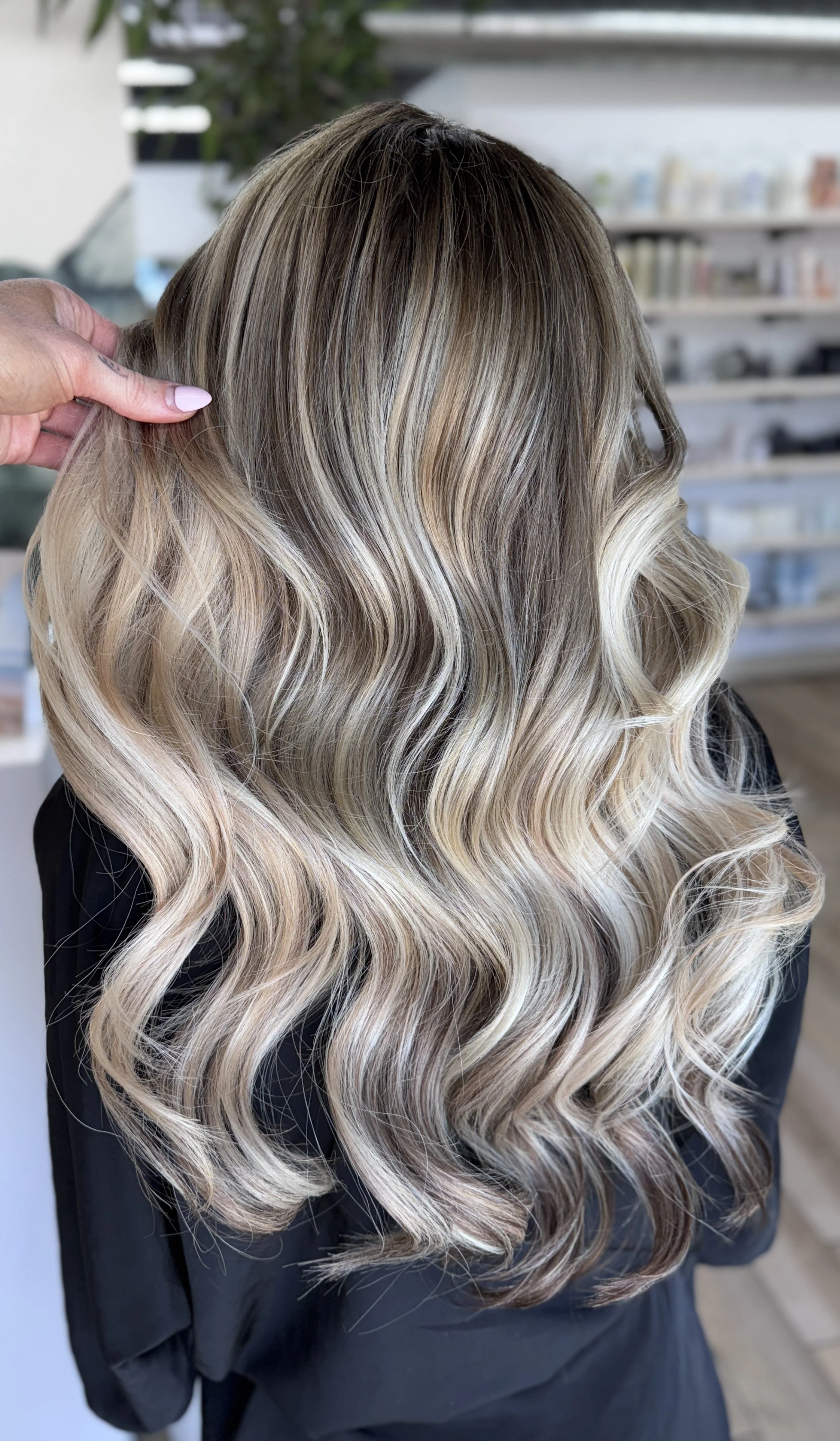 A woman with long, wavy blonde hair with darker roots and lighter highlights, styled in loose curls.