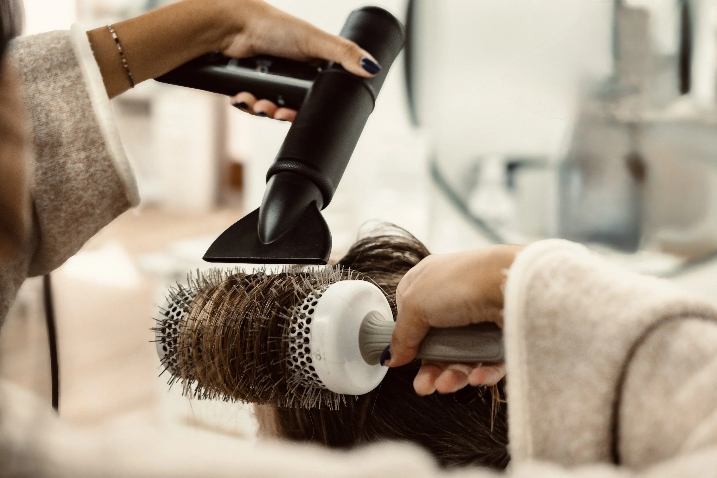 Someone uses a heated air tool with a round brush attachment to style a person's brown hair.
