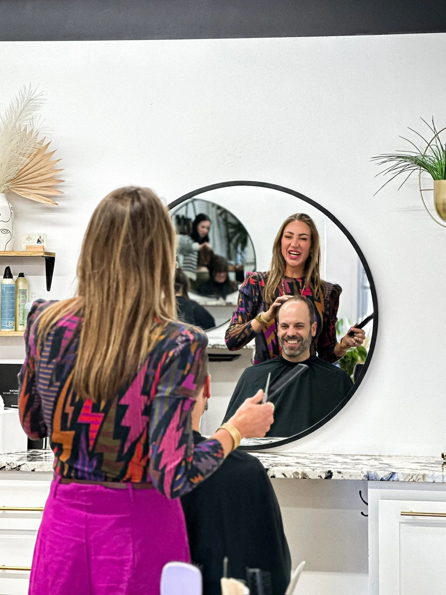 A woman in a colorful zigzag-patterned jacket and fuchsia skirt is giving haircuts to a man in a black cape, reflected in a round mirror in a modern salon.