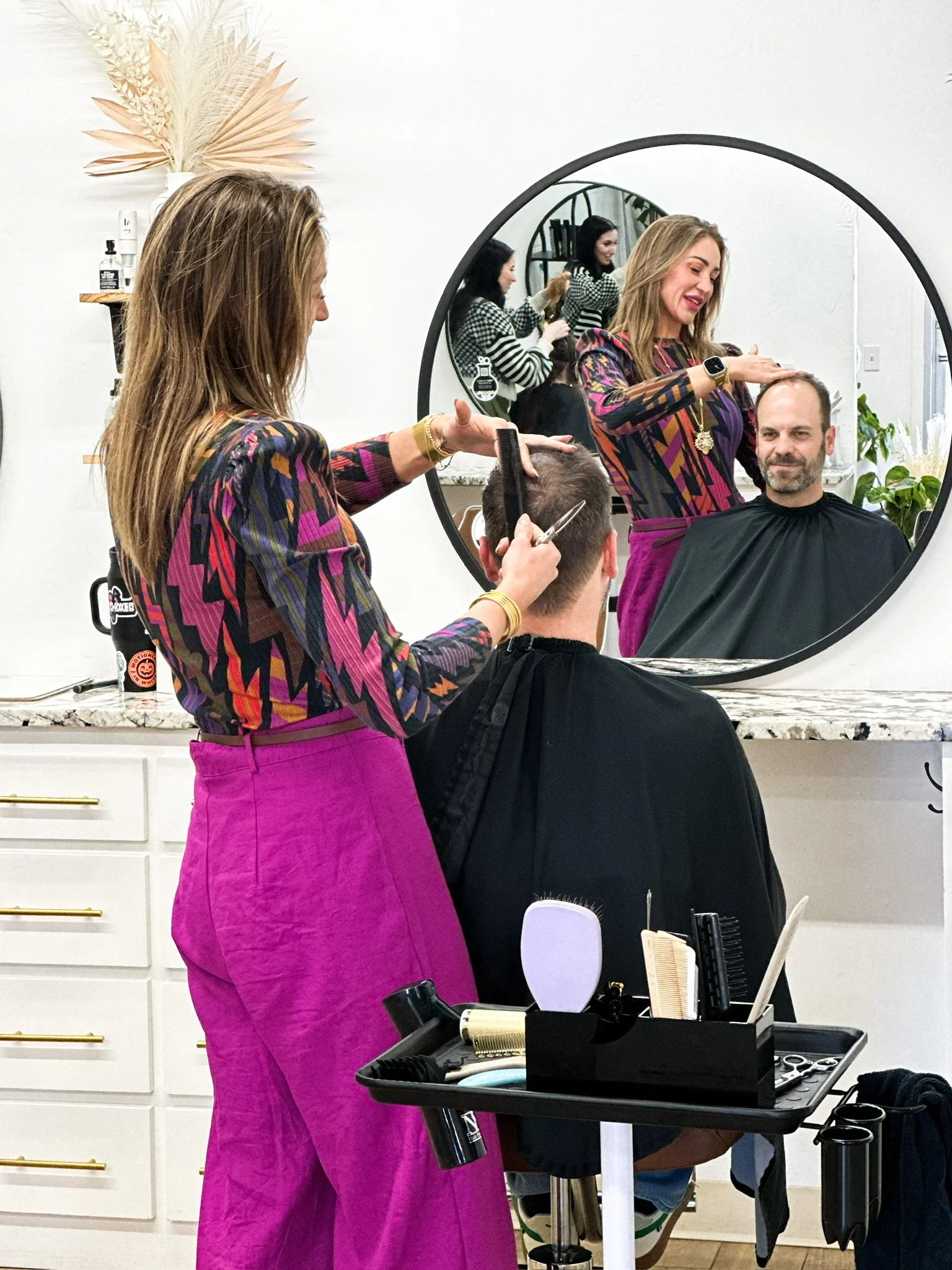A hairstylist cuts a man's hair in a salon, seen through a mirror that shows both the hairstylist and man, with the stylist wearing a colorful patterned top and bright pink pants.