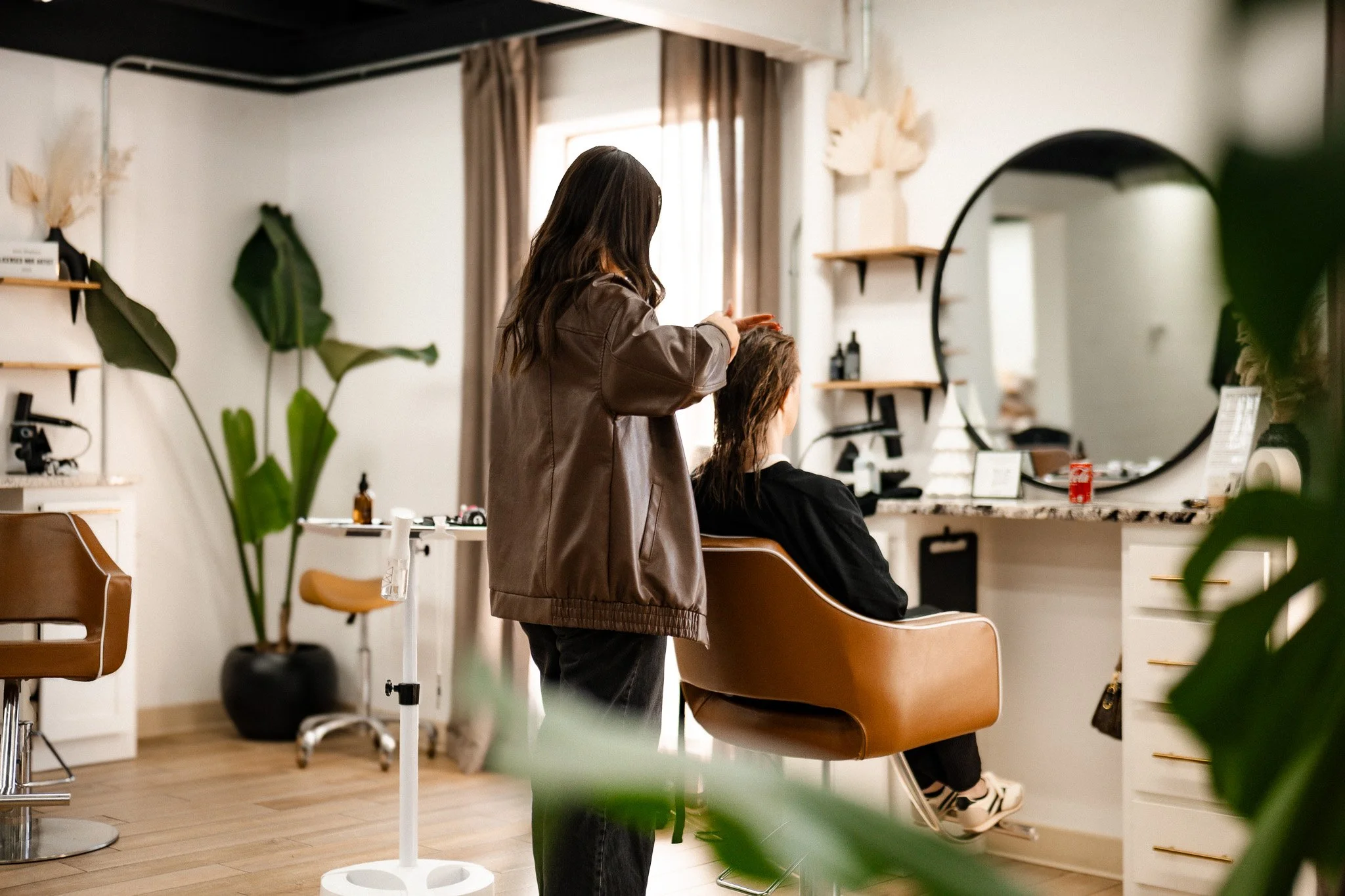 A hairstylist is drying and styling a woman's hair in a modern, well-lit salon with a large round mirror, purple curtains, and plants.
