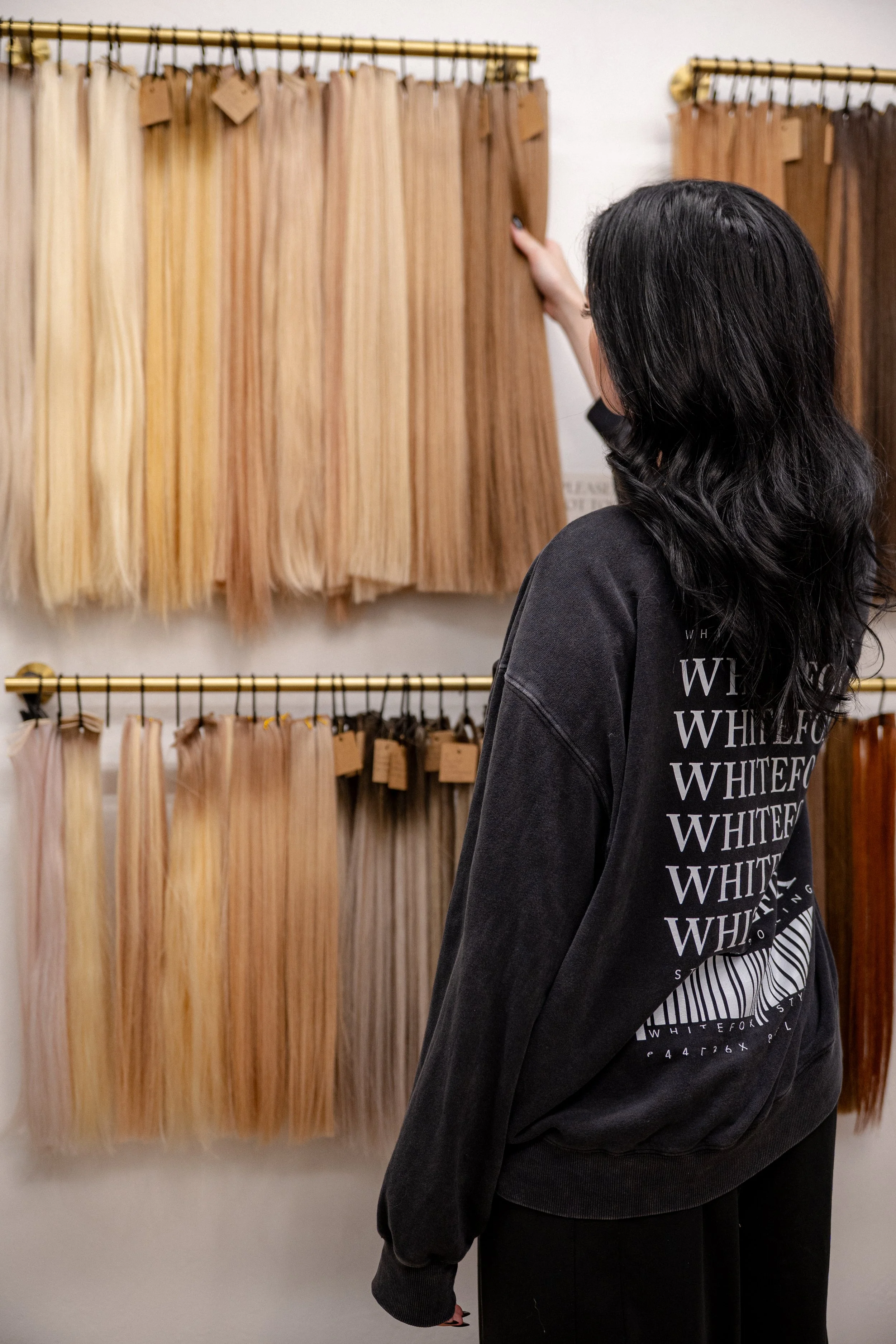 A woman shopping for hair extensions or wigs at a store, looking at hanging hair extension samples in various shades of blonde and brunette.
