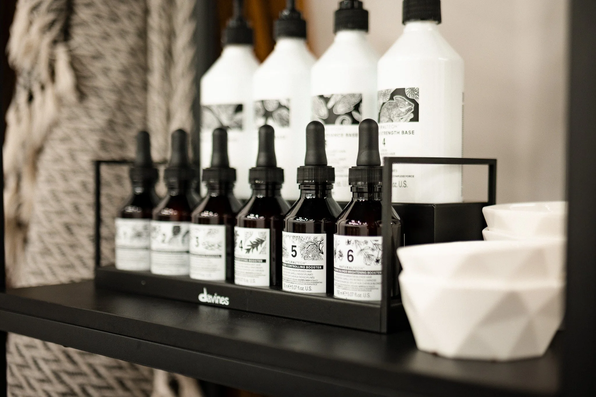 A shelf holding several small dark glass bottles with dropper caps, labeled with botanical illustrations and numbers, and a white rectangular tray with white bowls beside it.