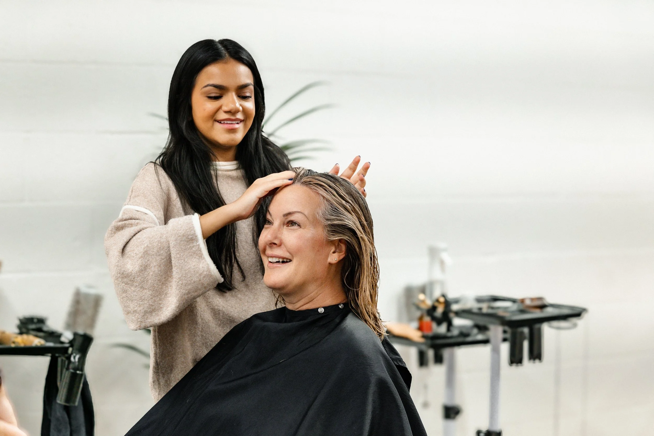 A hairstylist is smiling and massaging the scalp of a woman sitting in a salon chair, with hair in a cape, in a hair salon.