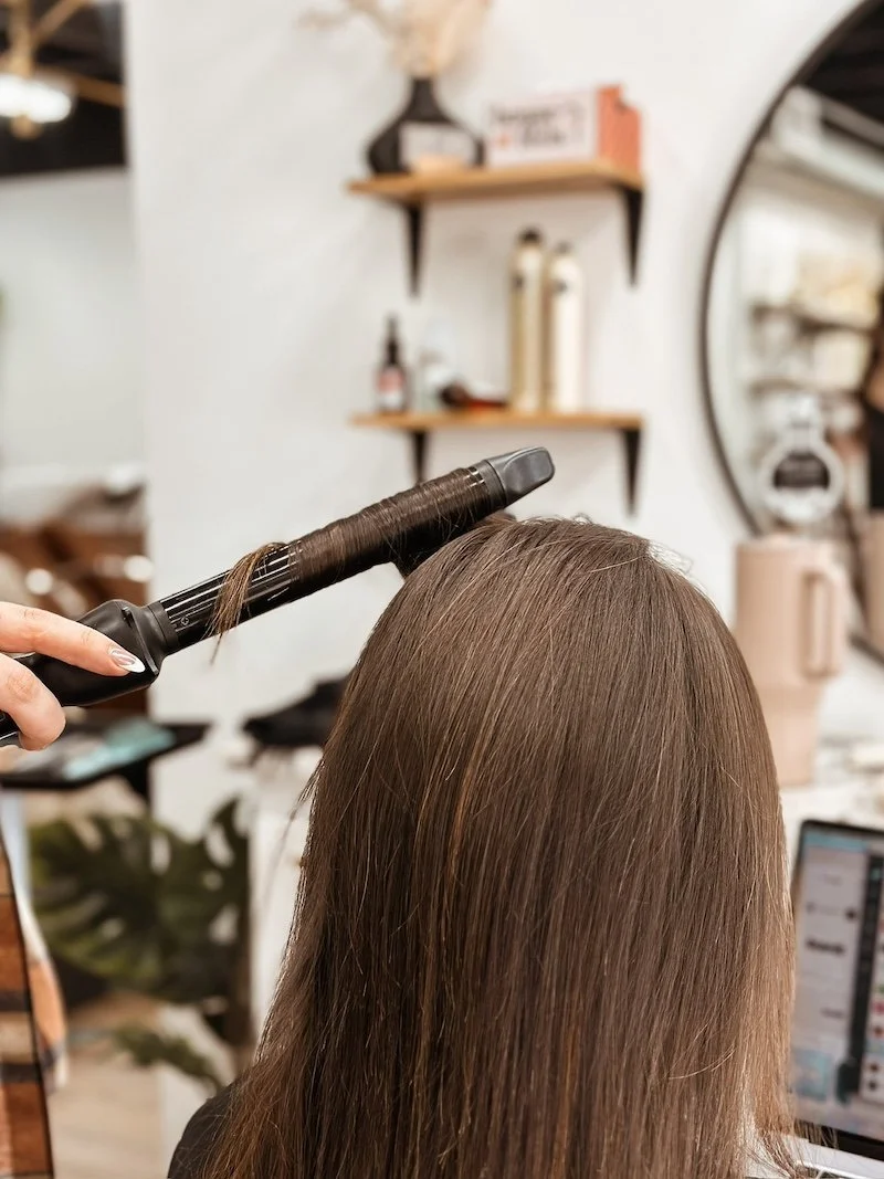 Person styling long brown hair with a curling iron in a salon.