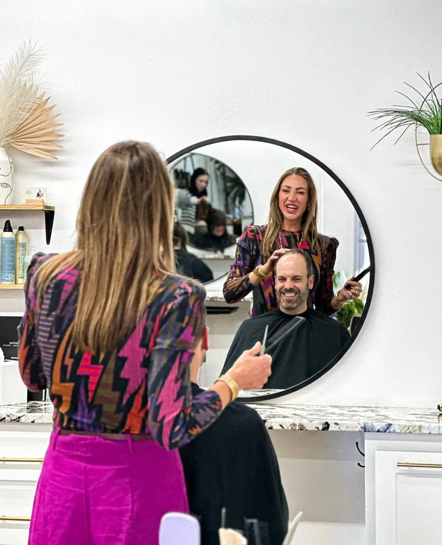 A woman getting her hair styled by a hairstylist in a salon, with both women smiling and a man getting a haircut reflected in a round mirror.