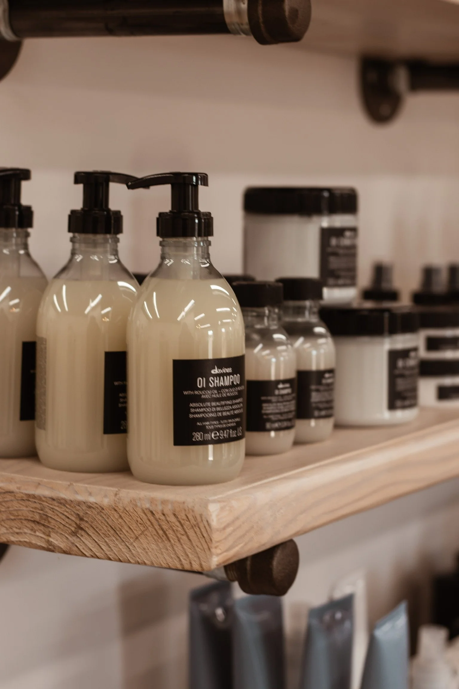 Three bottles of Davines Oli Shampoo on a wooden shelf in a salon or store, with other bottles and containers in the background.