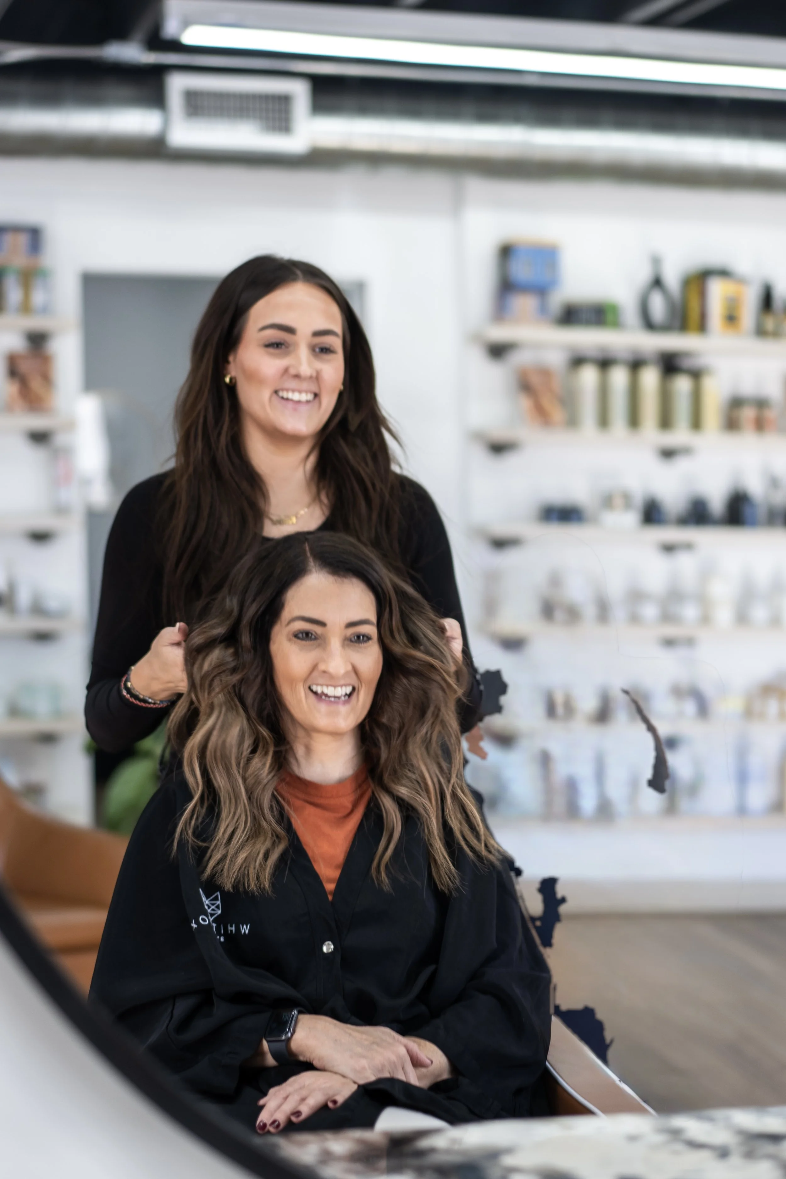 A woman with wavy, shoulder-length hair is sitting in a salon chair, smiling, while a hairstylist with long dark hair stands behind her, smiling and preparing to style her hair. The salon has shelves with various hair and beauty products in the background.