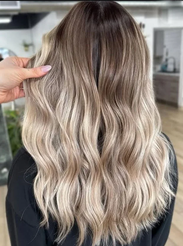 Back of a woman's head showing long, wavy blonde hair with darker roots, styled in loose waves.