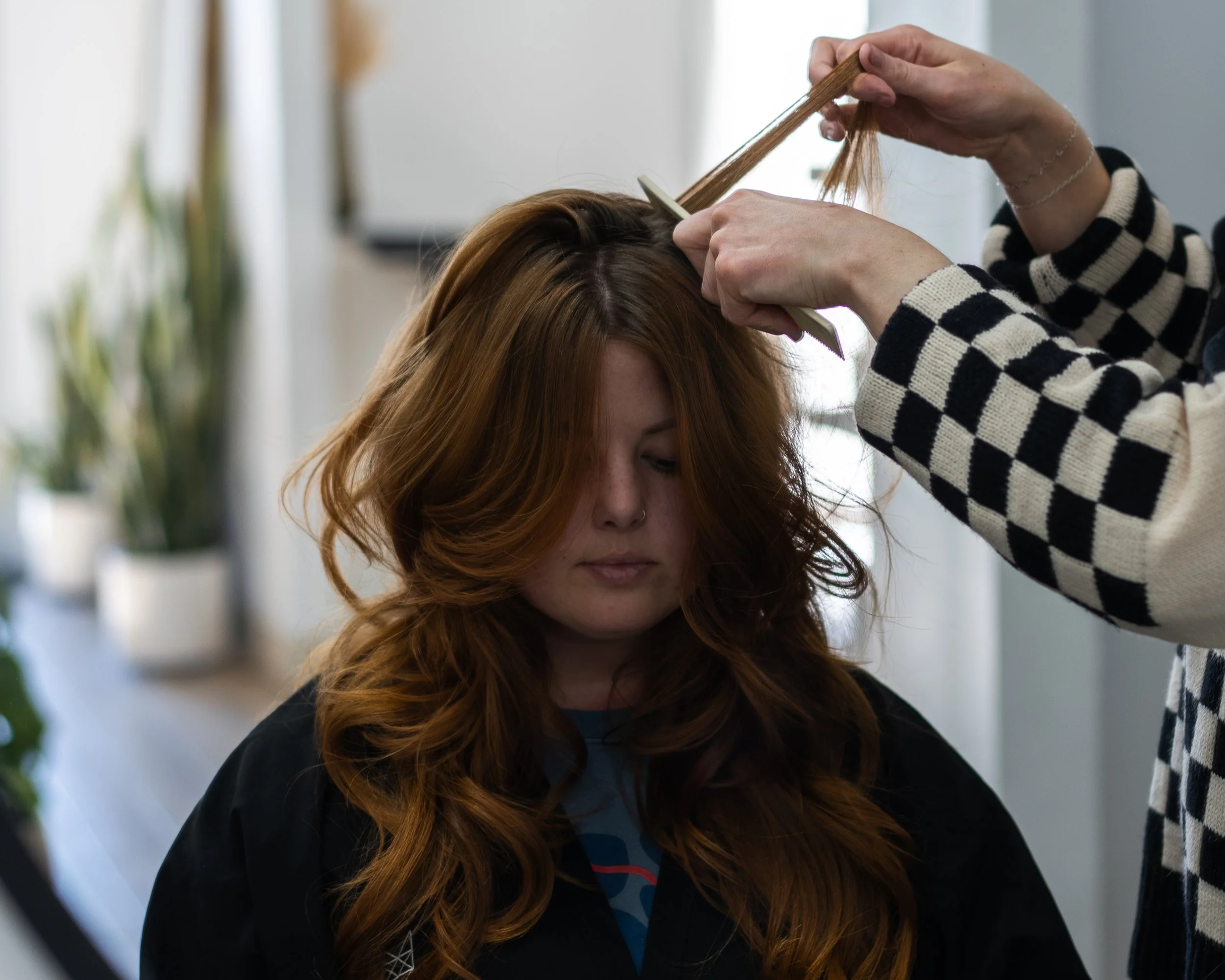A woman with long, wavy red hair sitting in a hair salon, getting her hair styled by a stylist. The stylist is holding a section of hair and a comb, working on her hair.
