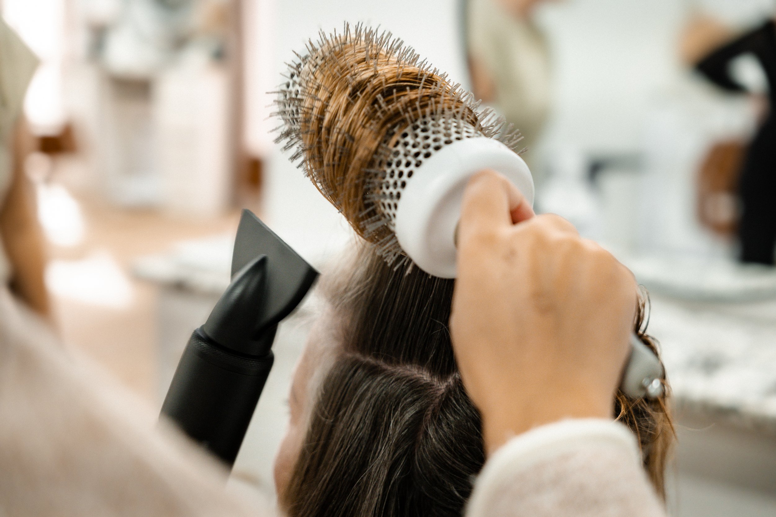 Person styling hair with a round brush and a blow dryer in a salon.