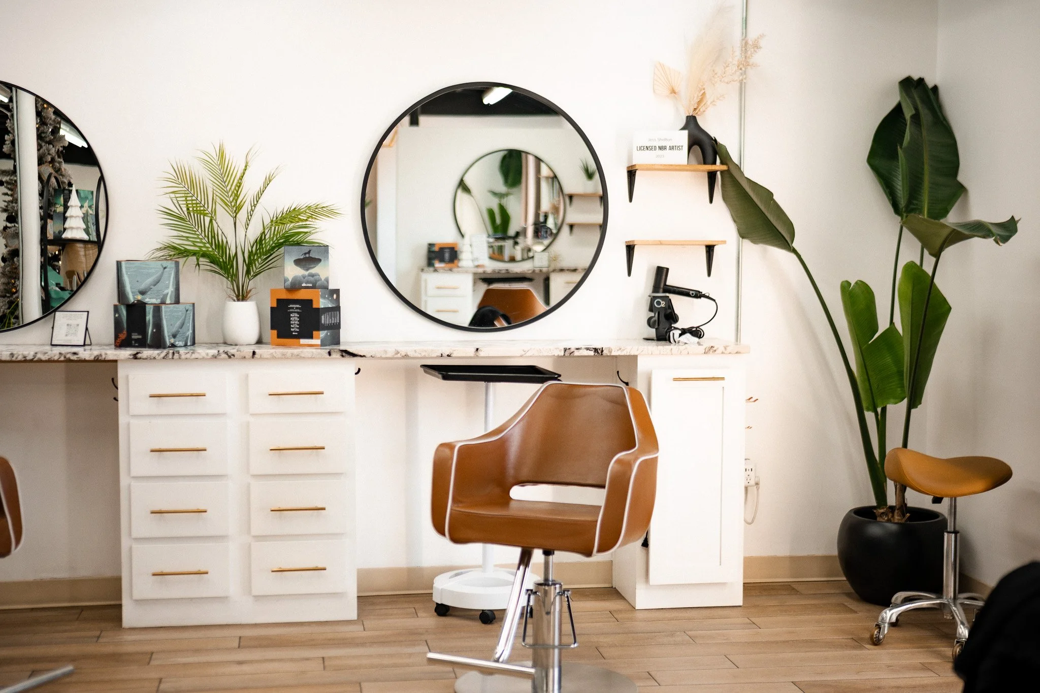 Interior of a hair salon with two round mirrors, a white marble counter, a brown salon chair, green plants, and wooden shelves.