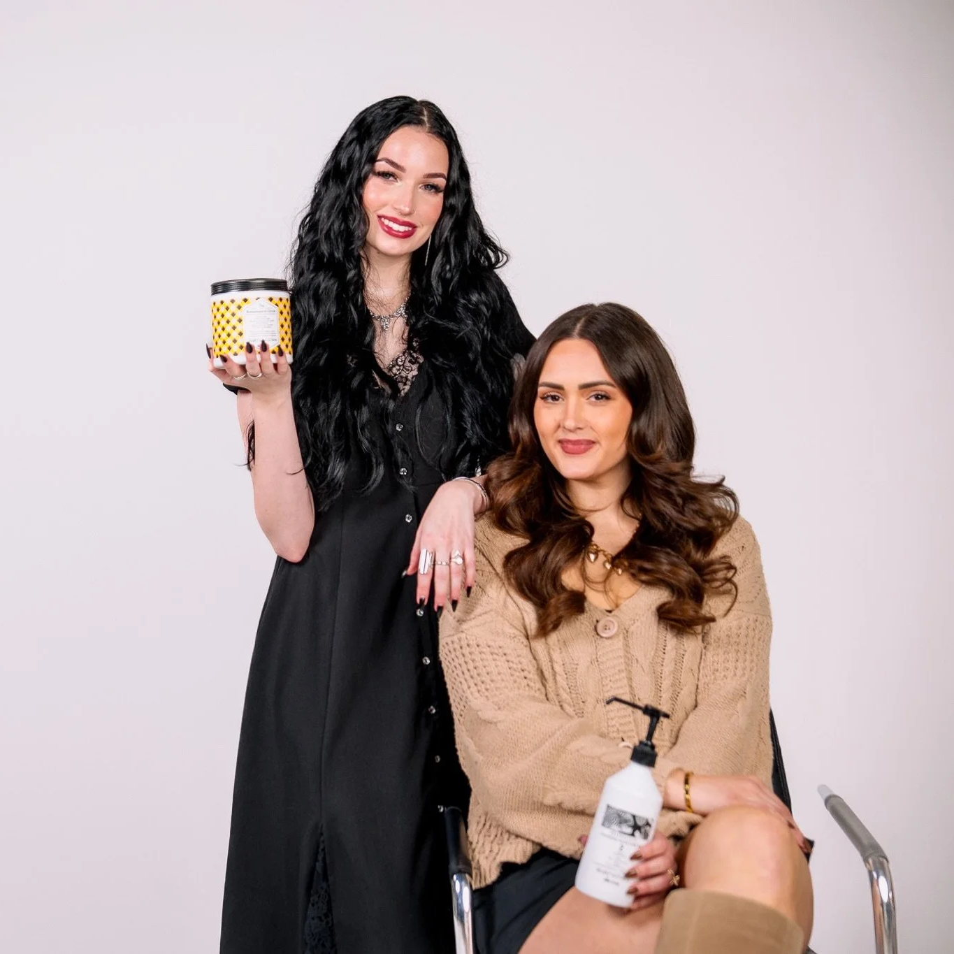 Two women, one sitting in a wheelchair with a brown sweater, holding a white bottle, and the other standing behind her with long black wavy hair, wearing a black dress, holding a yellow and black container, both smiling.