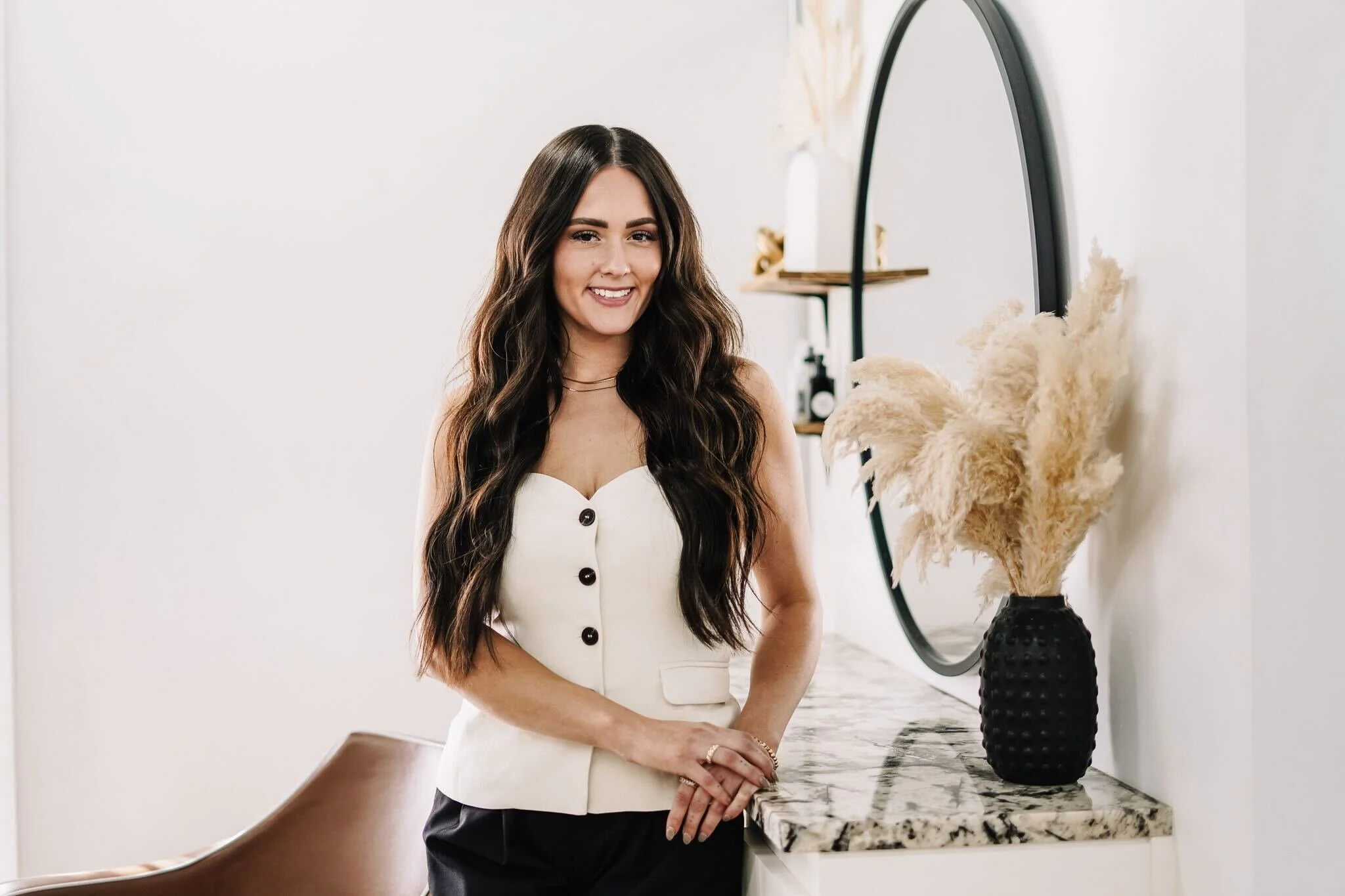 A woman with long, wavy brown hair standing indoors next to a black vase with dried pampas grass on a marble-topped table, smiling at the camera.