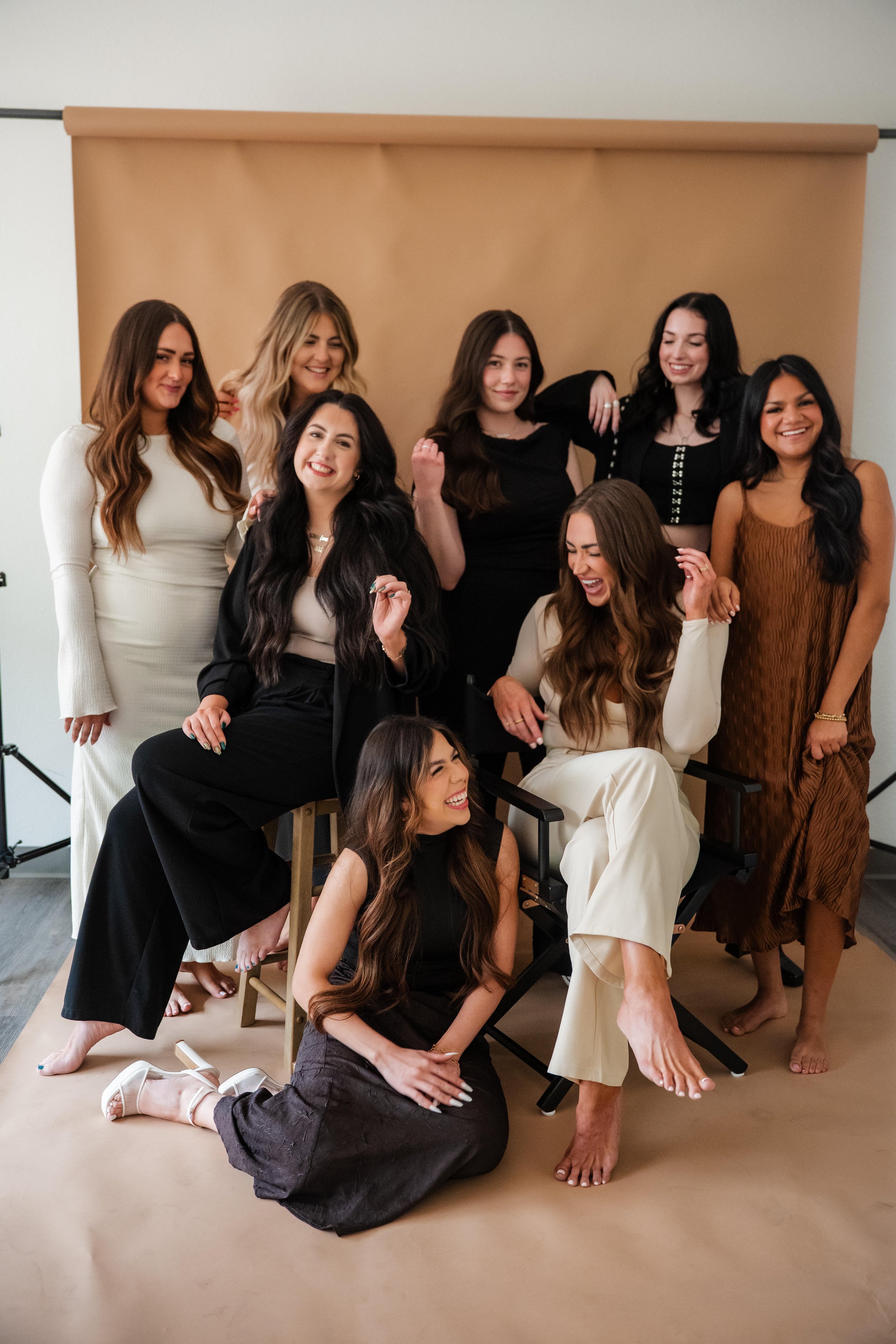 Group of nine women posing and laughing in front of a beige backdrop during a photoshoot.