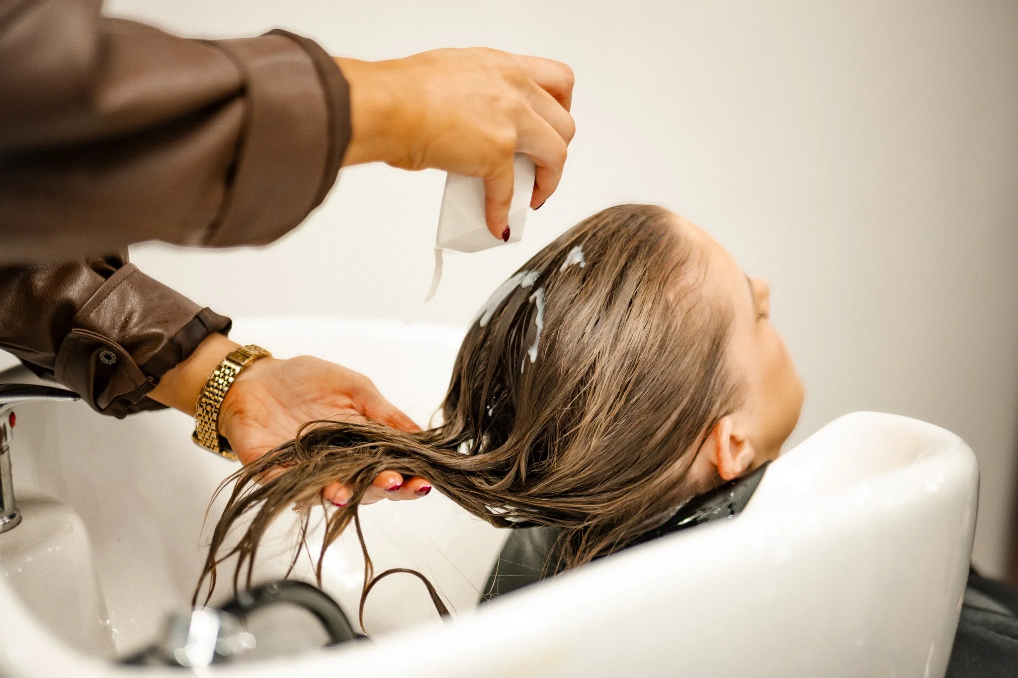 A person is washing a woman's wet hair in a salon sink, using a shampoo bottle.